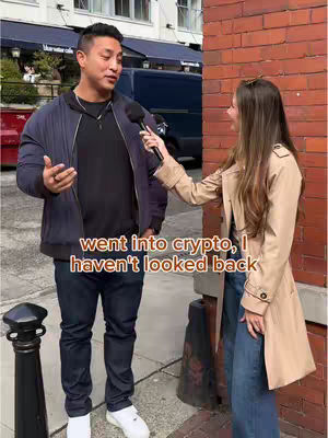 A man is being interviewed on the street by a woman. The man is wearing dark clothes and and has his hand gesturing, looking and talking at the woman. The interviewer is facing the man, holding a microphone and is wearing a brown coat. Both of them are standing against a brick wall as a street scene.