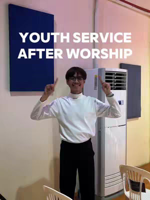 A young man wearing a white turtleneck top and black pants stands indoors. He is smiling and points his index fingers upwards with both hands. Behind him, a large blue panel and an air conditioning unit are visible. Text overlayed says, 'YOUTH SERVICE AFTER WORSHIP' with a white font.