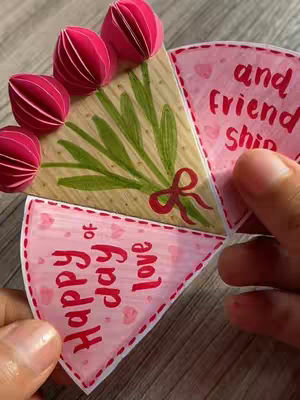 The cover image shows a handmade Valentine's Day card in the shape of a tulip bouquet. Four pink paper tulips are placed on top of a central triangle with a painted green leaves and a red bow. The card is composed of multiple triangular sections with handwritten messages in red, held casually by a pair of hands.