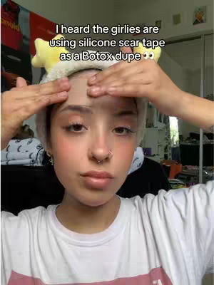 A selfie-style shot features a person with fair skin, wearing a white t-shirt with a pink stripe. They have light makeup, a nose ring, and a headband with yellow star-shaped decorations. The person's hands are raised to the forehead, where a patch of tape is applied, against a cluttered domestic setting.