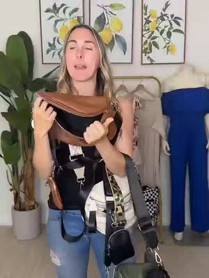 A woman stands in front of a wall, holding and displaying multiple bags of various styles and colors. She is wearing a tank top and jeans. The background includes framed artwork, a plant, and clothing racks.
