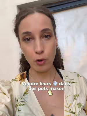 A woman with brown curly hair and hoop earrings is looking at the camera and talking. She is wearing a floral top and a gold necklace. Text overlayed on the image reads, "Vendre leurs dans des pots mason". The background is a blurry room interior.