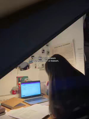 A person studies at a desk in a dimly lit room. A laptop and open books are on the desk. The person is seen from the back.