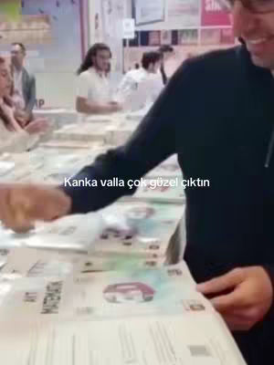 A man is holding and flipping through a book at a table. Other people are visible in the background in a brightly lit area. The overlay text reads, "Kanka valla çok güzel çıktın".