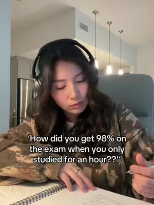A woman with long dark hair wearing headphones is looking down at a notebook on a desk. She is wearing a patterned jacket and a ring on her finger. The background includes overhead lights and part of a kitchen.