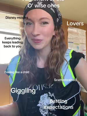 A woman with long, wavy auburn hair and fair skin looks directly at the camera with a slight smile. She wears a black headscarf patterned with black and white designs, a black t-shirt containing white graphic elements over a lighter blue sleeveless top with reflective yellow-green trim. Around her are text overlays with words and phrases.