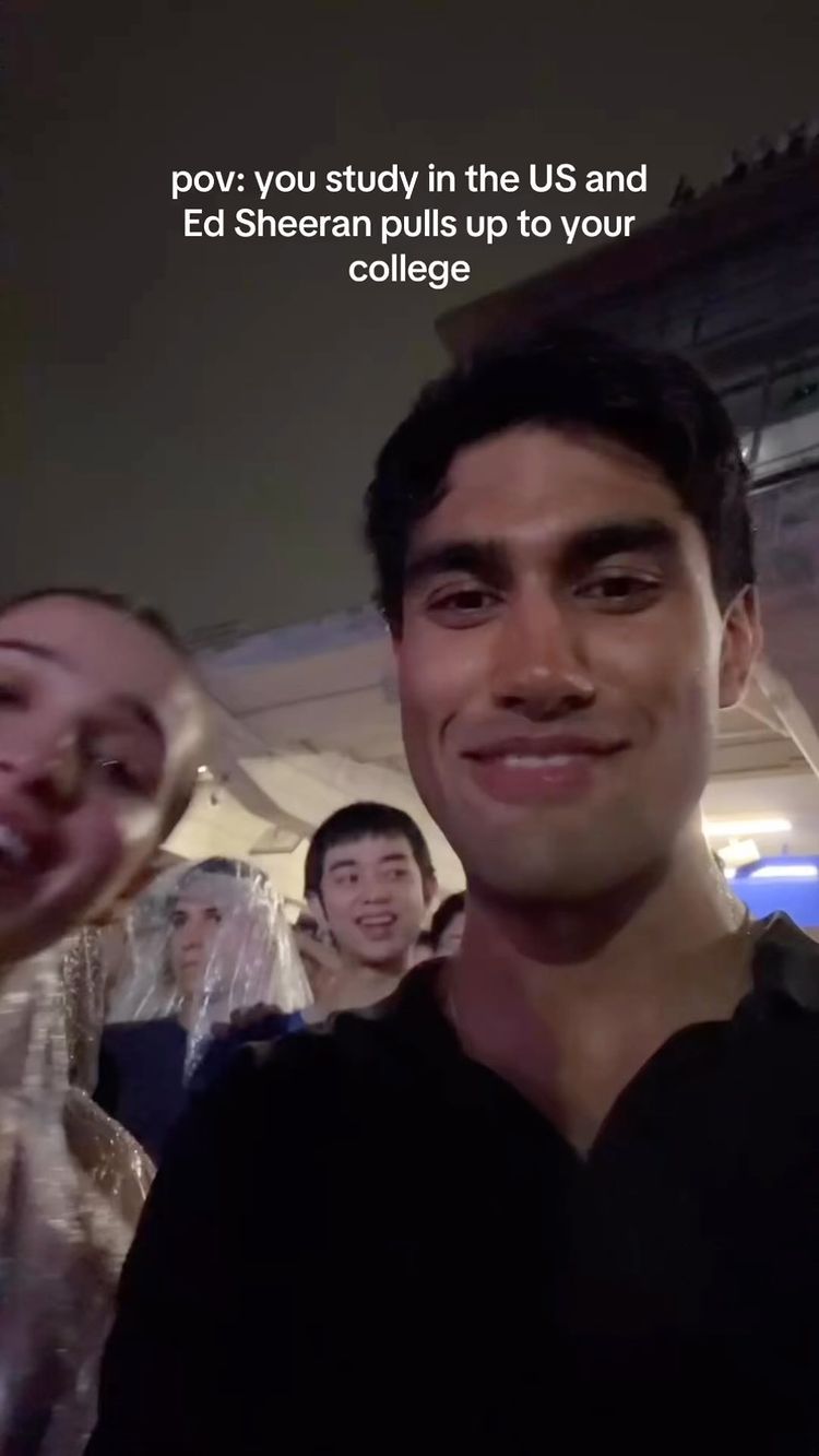 A selfie-style shot featuring a man smiling widely, with two other people in the background. The man is in the foreground, taking up most of the frame, with fair skin and dark hair. Behind him, slightly out of focus, are two other people. The background is blurred with a few visible lights, suggesting an outdoor setting at night. The composition is casual and the lighting seems to be artificial.