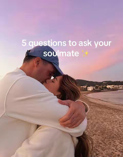 A couple is embracing on a beach at sunset. The man is wearing a blue cap and a white sweatshirt and is holding the woman close. The woman has long brown her and is also wearing white cloth. The background has pink and purple shades, with water and trees in the distance.