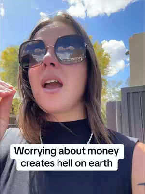A woman with sunglasses looks up with an open mouth set in front of a sky and partial building. The overlayed text reads, 'Worrying about money creates hell on earth'.