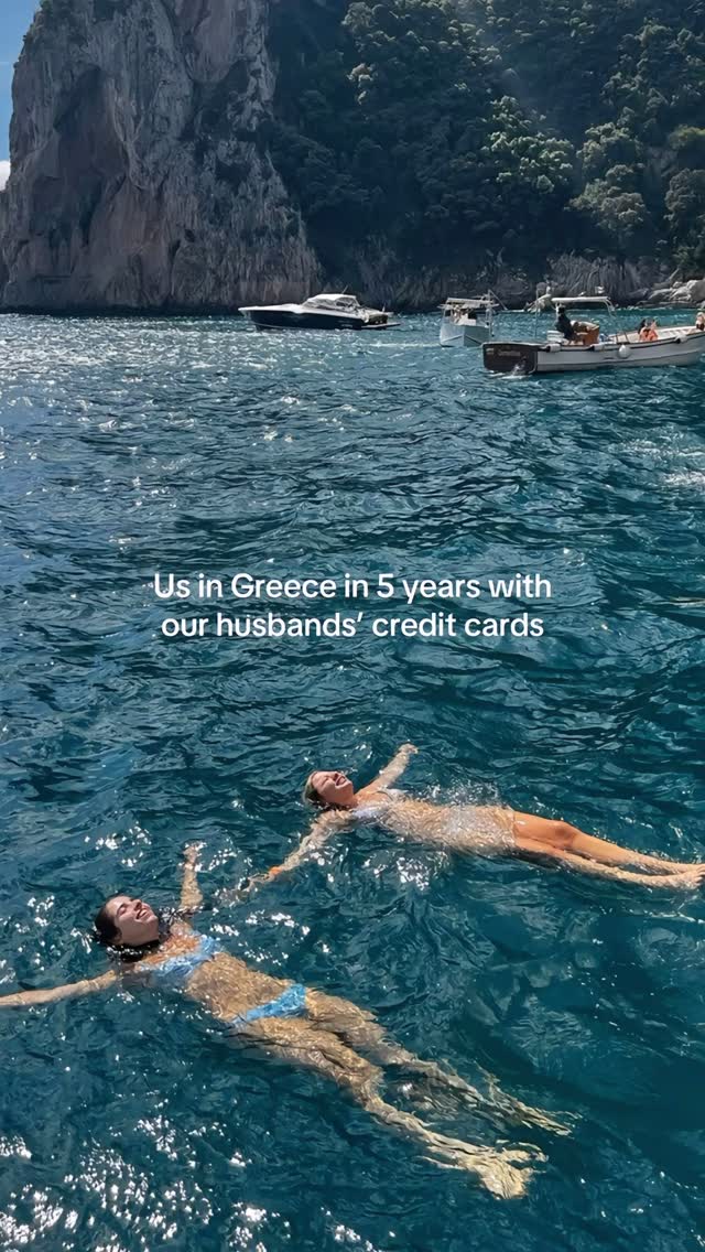Two women floating in the water with their arms outstretched. The water is clear blue, and the sky is sunny and bright. There are boats and mountains in the background.