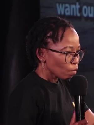 A woman with short, dark hair wearing glasses and a black shirt speaks into a microphone. The background is dimly lit, with some indistinct text visible above her.