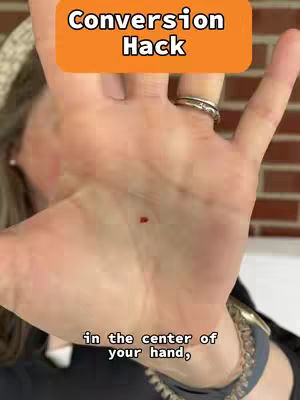 A well-lit, close-up shot centers on a person's open palm, with focus given to a red dot on the skin. The hand is in the foreground, fingers spread slightly apart towards the top. The background includes elements like a brick wall and a partial view of an arm with a watch. Additionally, the top of the image has an orange rectangular banner with white text.