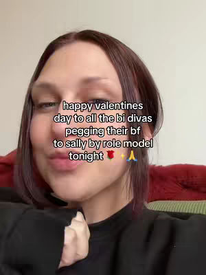 A selfie-style shot featuring a person with fair skin and dark maroon hair. They are wearing a black top, and their hand is touching their face. The text overlayed on the image reads: "happy valentines day to all the bi divas pegging their bf to sally by role model tonight."