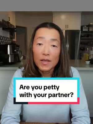 A woman with brown hair and fair skin is centered in the frame. She is positioned in front of a domestic background (kitchen or dining room). The background is somewhat out of focus, with light-colored cabinetry, parts of a stainless steel appliance, and a glimpse of a doorway. The woman is wearing a light-colored top. Overlaying her is a colorful text box in a rectangular shape, which reads "Are you petty with your partner?"