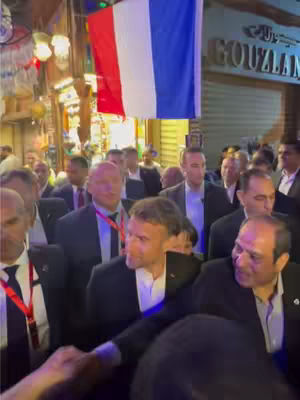 A crowd of people, mostly men in suits, gather in a public outdoor setting. A large French flag hangs above the crowd, and a sign reading "GOUZLAN" is visible in the background. The image is brightly lit.