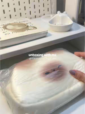 The image shows a shelf with items for an unboxing video setup. A white shelf holds a stack of books with a decorative cup visible, and a small white bowl. Below, a square package wrapped in plastic is the central focus, slightly obscured by the wrapping.