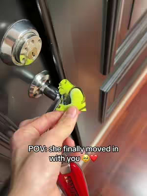 The image shows a hand holding a small cutout of Shrek next to a door with a key in the lock. The background appears to be a modern apartment or home.