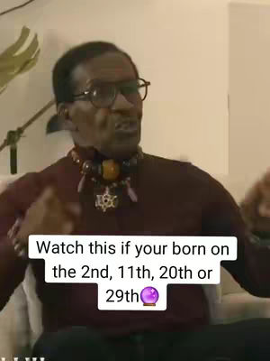 The image features a middle-aged Black man framed from the chest up, sitting on a beige sofa. He wears glasses and a maroon long-sleeved shirt. His neck is adorned with a beaded necklace and necklace with pendant; his expression, animated and serious, suggests he's speaking directly to the viewer. A white text box at the bottom of the image provides the hook. The lighting is soft.