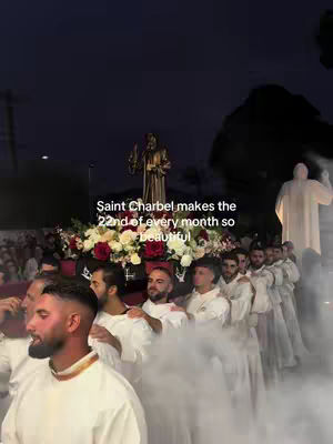 The image shows a procession at night, possibly a religious event. In the foreground, several men in white robes are visible; one of them, in a close-up, appears to be the main subject holding a necklace. In the mid-ground, a statue on a decorated float carried by people is surrounded by flowers and lights. Behind the float are more people dressed in similar white robes, suggesting a formal procession. The background is dark with other structures and some figures that appear like individuals. The image is in a vertical format, and the overall atmosphere is solemn and somewhat atmospheric due to the darkness and the use of lights.