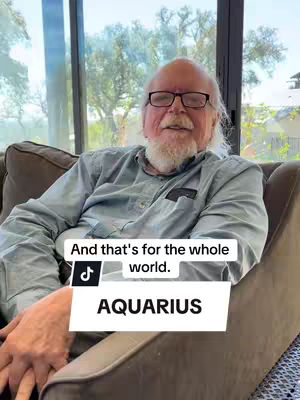 An older man with a white beard and glasses is sitting in a brown chair in front of a large window partially visible behind him. There is a white text box at the bottom with the word "AQUARIUS".