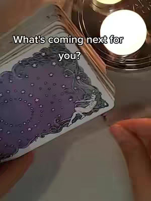 The image shows a close-up view of a tarot card deck being held next to a lit candle. The deck has a purple and white design with stars and a figure. Beneath the deck are the edges of a table and near the top right, there is a lit candle.