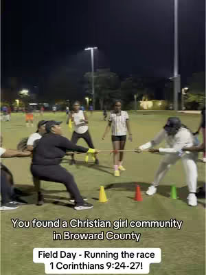 A nighttime field day with a group of young women playing tug-of-war. The image is in a dark setting with stadium lights illuminating the scene and the participants. Text overlays describe this is a Christian girl community in Broward County, and mentions 'Field Day - Running the race, 1 Corinthians 9:24-27!'