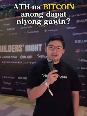 A man in glasses holding a microphone and speaking to the camera, with a text overlay. Behind him is a backdrop with logos and text.