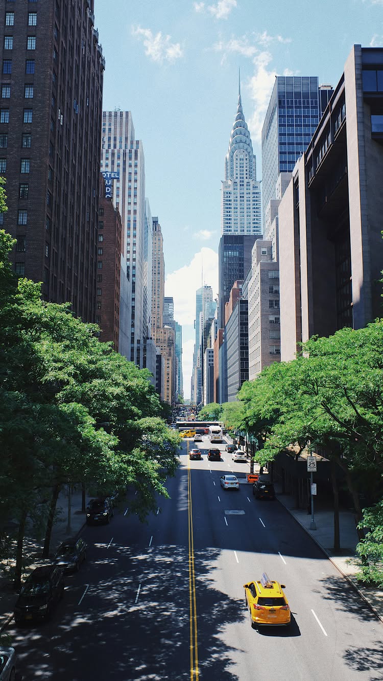 A scenic, eye-level shot of a New York City street, framed by tall buildings. A road stretches into the distance, with a golden-yellow taxi in the foreground. The Chrysler Building is a focal point in the background, under a blue sky with scattered clouds.