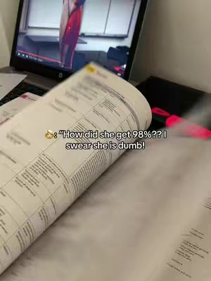The image shows a laptop open with a video playing on it, a book open in the foreground, and a text overlay that says, "How did she get 98%?? I swear she is dumb!". The composition is a cluttered scene with a focus on study materials combined with online resources, suggesting a study-related theme.