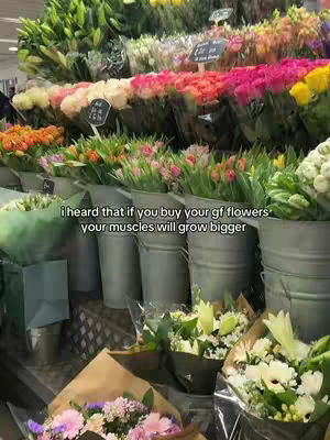 The image shows a flower shop with various bouquets and buckets of flowers. The foreground features bouquets wrapped in brown paper, while the background displays flower arrangements in metal buckets. Text overlay adds a humorous line about buying flowers for muscle growth.