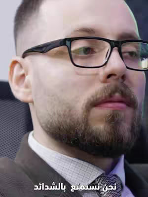 The cover image is a close-up shot of a man wearing black-framed glasses and a collared shirt with a patterned tie. The man has short, neatly styled dark hair and a well-groomed beard. The background is a blurry indoor setting with dim lighting. The man is looking slightly off-camera.