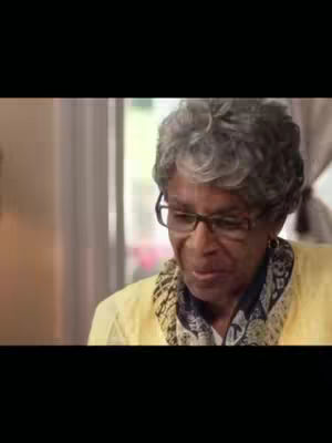 The image shows an elderly Black woman wearing glasses and a yellow top, looking down and to the side. The background is softly blurred, suggesting an indoor setting.