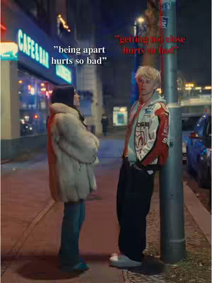 A nighttime street scene with two people, a man and a woman, standing close together. The man leans against a utility pole, wearing a jacket, while the woman stands facing him, wrapped in a large coat. Text overlays are added to the image, positioned near each person, with each one having quotes. The background includes a brightly lit cafe or bar, its sign visible, and other buildings illuminated by city lights. The overall tone is cinematic, suggesting a dramatic or romantic interaction.