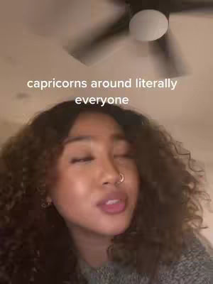 A close-up shot of a woman with curly hair. The background shows a ceiling fan. Text overlay says "capricorns around literally everyone."