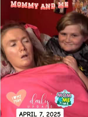 A selfie-style shot of a woman and a young boy. The woman is in the foreground, partially visible, with a pink shirt, appearing to look unwell. Behind her and to her right is a boy, smiling and looking at the camera. The background includes a red wall.