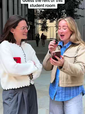 Two young women are standing outside, facing each other with a blurred background, one is holding a phone, and the other one seems to be holding a small microphone. Text in a white box at the top of the image, and the background depicts an urban environment.
