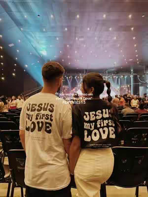 Two people, facing away, stand in a large open space that resembles a church or auditorium with a high ceiling and numerous lights. Both are wearing matching black t-shirts. The person in the foreground is wearing a white shirt that says 'JESUS my first LOVE.' The background shows a blurred crowd and stage.