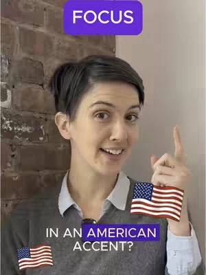 A woman with dark hair and a grey sweater is centered in the frame. Behind her is a red brick wall and a white background. A purple rectangular overlay with white text saying 'FOCUS' is situated at the top of the frame. Two flags are visible, one at the bottom right and another at the bottom left. Another purple rectangular overlay with white text saying 'IN AN AMERICAN ACCENT?' is overlayed near the bottom of the frame.