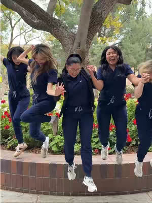 A group of five women in navy blue scrubs are jumping against a backdrop of a green tree. They are captured mid-air.