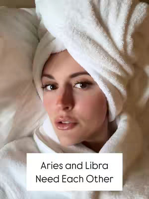 A woman with light skin lies on a bed, her head resting on a pillow. She wears a white towel wrapped around her head like a turban. The image has a soft, warm tone. A white rectangle at the bottom of the image has the text "Aries and Libra Need Each Other".