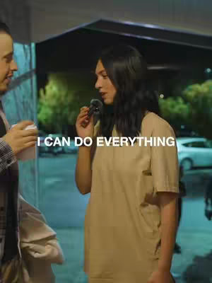 A woman holding a microphone and speaking, standing in front of a glass wall with a man on the left side, the background includes other people and cars. The main hook text is overlaid on the image.