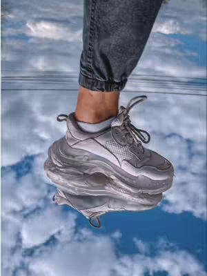 A person's leg and foot clad in gray athletic sneakers and dark gray jeans is reflected in a mirrored surface. The background features a sky filled with white clouds and patches of blue. The perspective is from below, looking up at the person's leg and shoe.