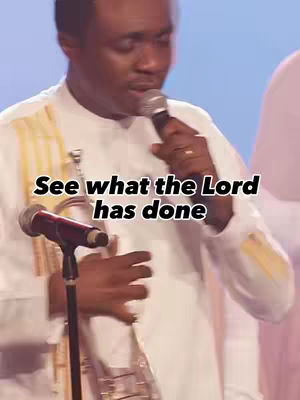 A man in a white shirt with gold accents is holding a microphone and singing in front of a medium blue background. Text overlay says, "See what the Lord has done," in a black font with a white outline.