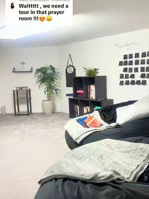 An interior shot of a room, possibly a prayer room, with a neutral color scheme. The room features a large plant in a pot, a cabinet with books and decorations, and a bed with pillows and blankets. The wall is white, and there are various decorative items. The lighting appears to be soft and ambient.