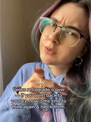 A selfie-style shot with a woman in glasses, a light blue hoodie, and colorful streaked hair. She points her index finger towards the viewer assertively. The background is a neutral, blurred space, keeping the focus on her expression and the text overlay. Text about Venus retrograde is displayed near her hand.