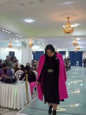 A woman in a black dress and a pink coat walks down a glass runway. A crowd is visible in the background. Formal and fashionable environment with chandeliers.