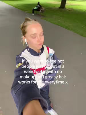 A woman in a dark blue and white jacket is shown facing the camera, taking a POV shot. In the background, a paved path leads towards a grassy area where a bench sits. Text overlay provides the content hook.