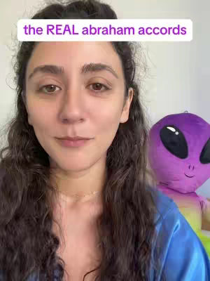 A woman with long, wavy brown hair is shown in a selfie-style shot. She wears a blue top and a delicate necklace. The background features a purple alien-like plushie and a white wall. Overlayed text reads 'the REAL abraham accords'.