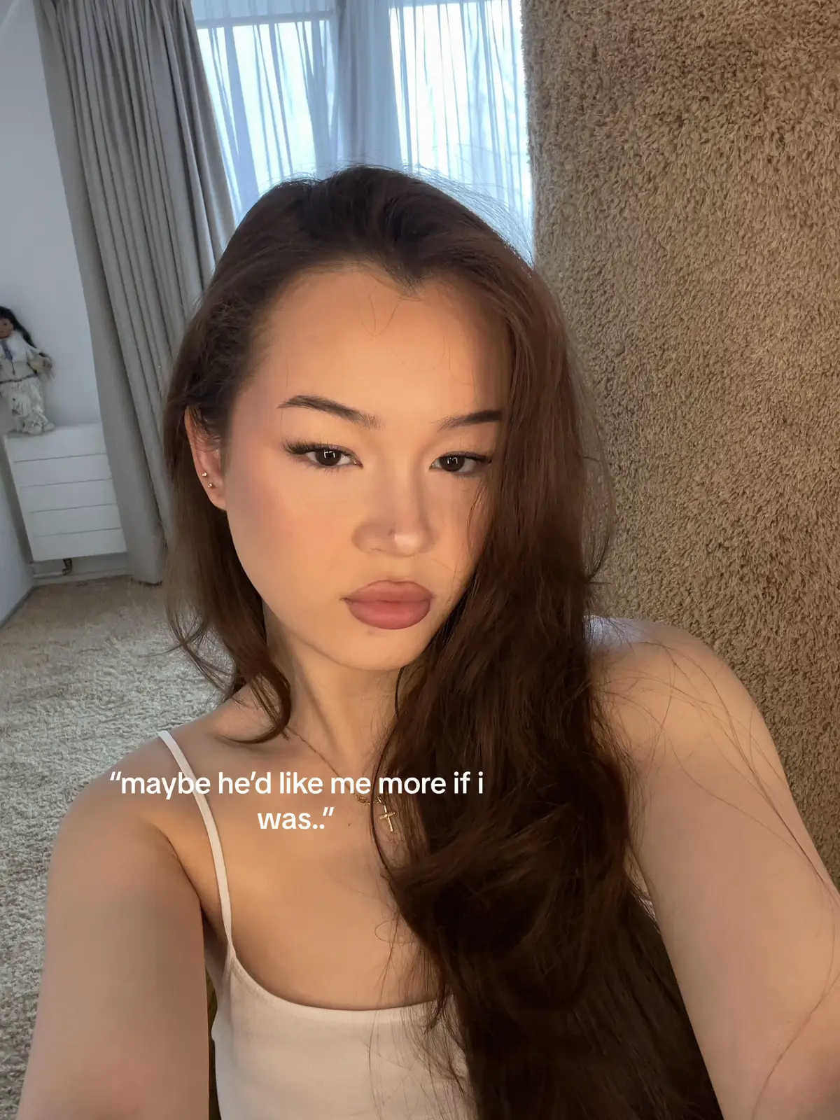 A selfie-style shot featuring a woman with long, brown hair and fair skin. She's wearing a white tank top and a delicate necklace with a cross pendant. The background is softly blurred, showing a neutral-toned carpet and wall. Text overlayed reads, "maybe he'd like me more if I was..".