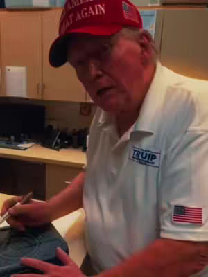 The image shows Donald Trump seated, appearing to write. He wears a red 'Make America Great Again' cap and a white shirt with the 'TRUMP' logo and a small American flag patch.